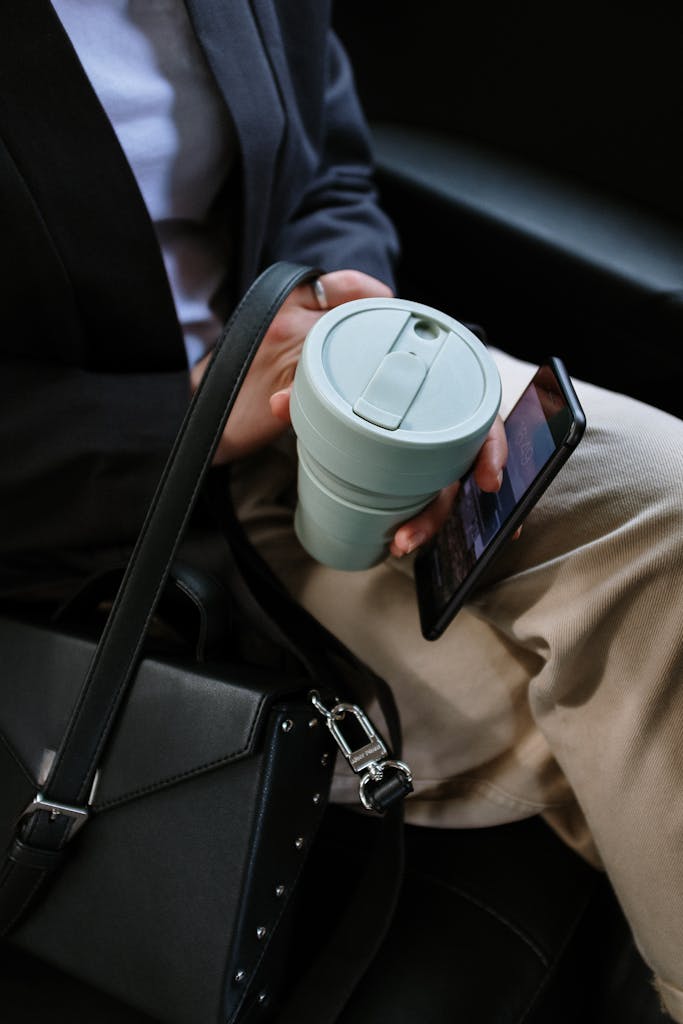 Person holding reusable coffee cup and smartphone in a car, depicting eco-friendly commute.