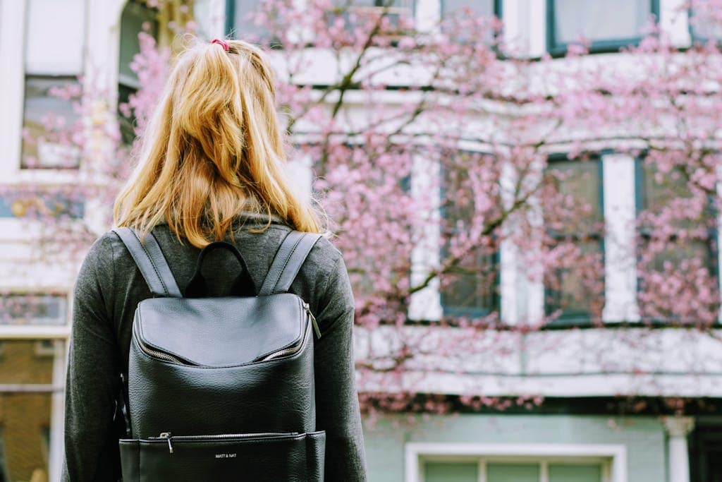 Blonde woman with a backpack stands by urban cherry blossom, hinting at travel and fashion.