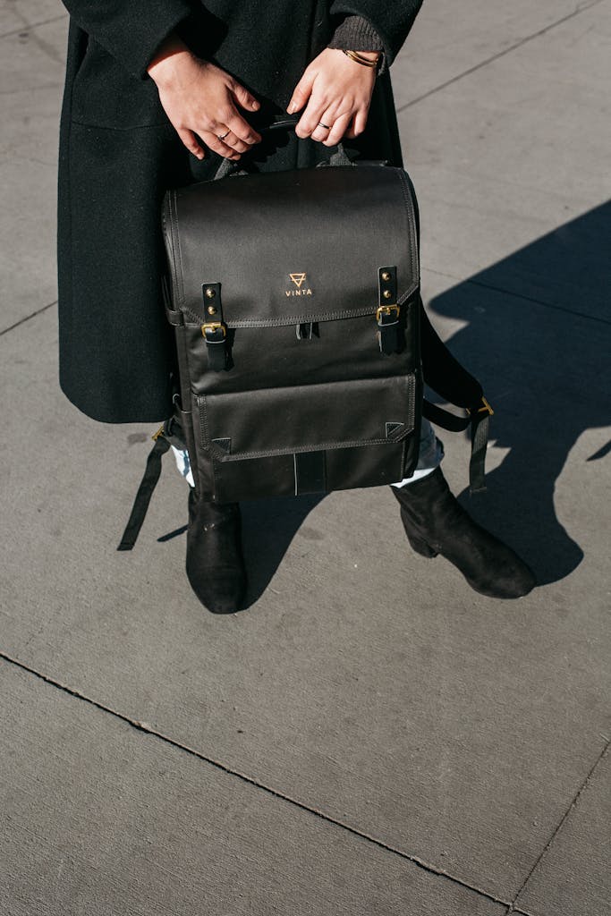 Fashionable black backpack held by a person on a city pavement in daylight.